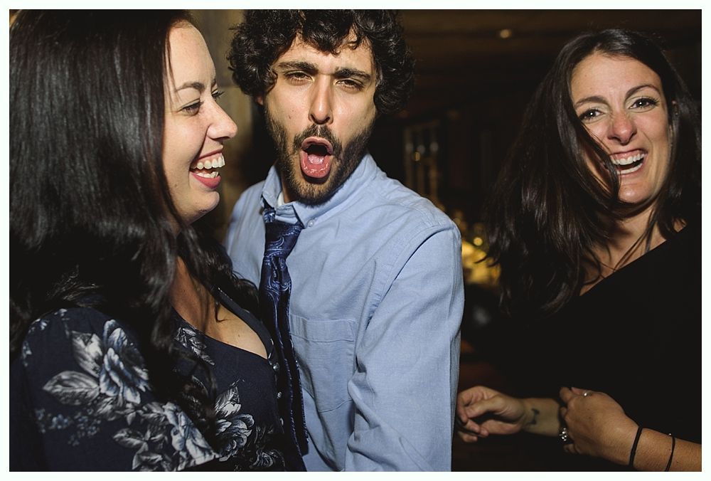 Three people laughing; a man with a surprised expression, and two women in a dimly lit setting.