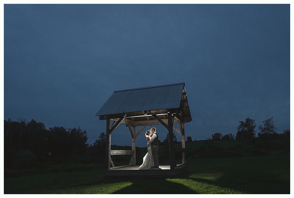 Couple embraces under a gazebo at dusk, illuminated by lights; field in background.