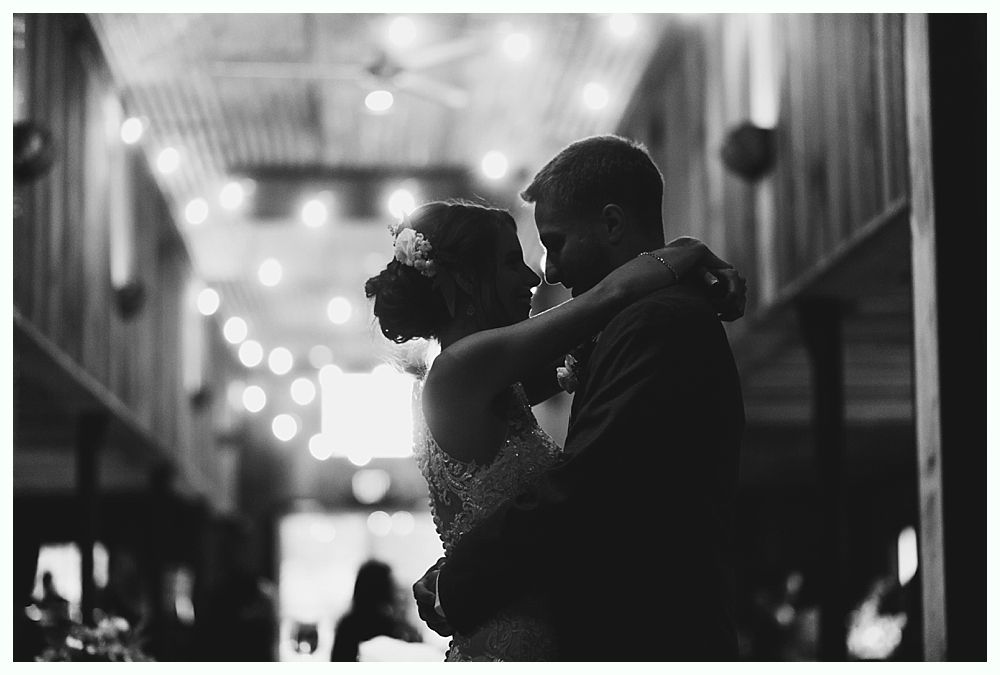 Couple dances closely in a dimly lit barn decorated with string lights.