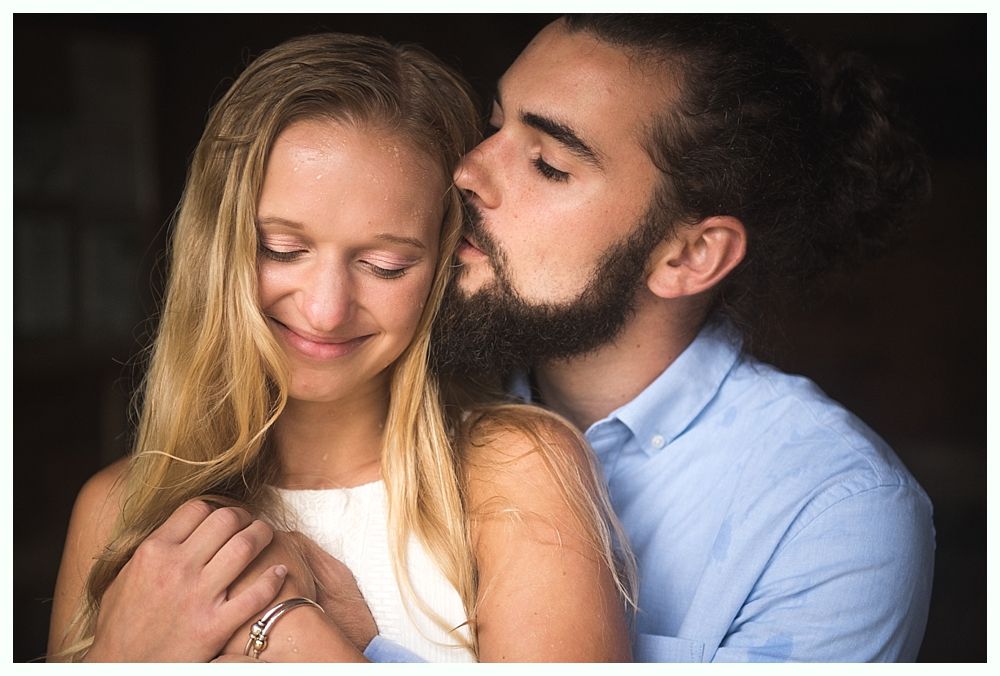 Man whispers in woman's ear, smiling. They embrace indoors; woman wears white dress, man has beard.