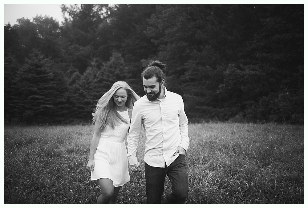Couple holding hands, walking through a grassy field. Trees in the background. Black and white photo.