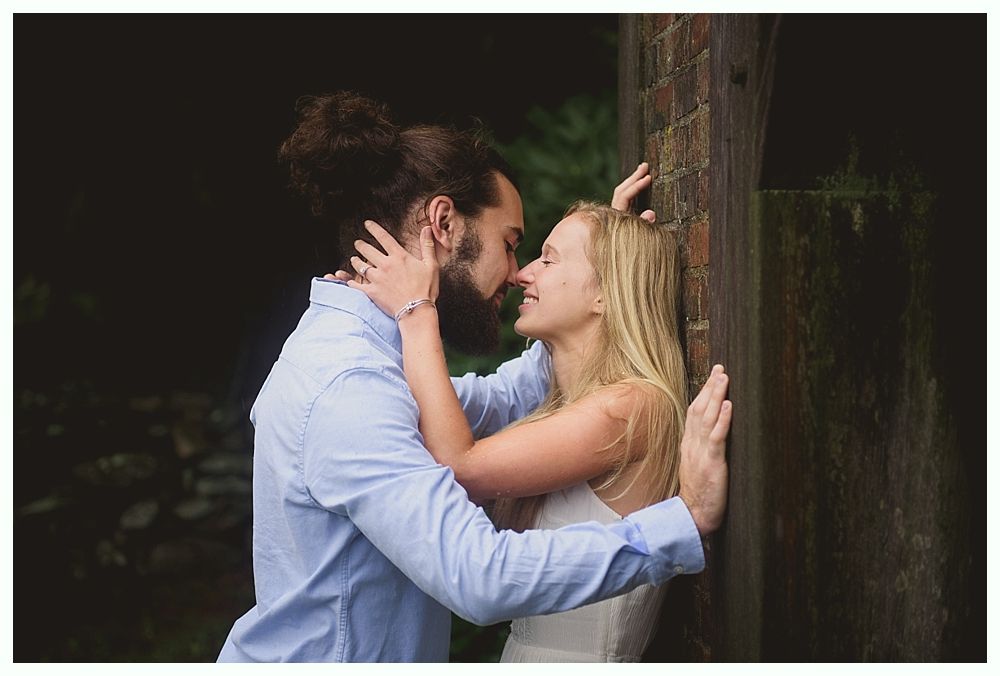 Couple embraces, leaning against a wooden structure; man with a bun, woman with blonde hair, dark background.