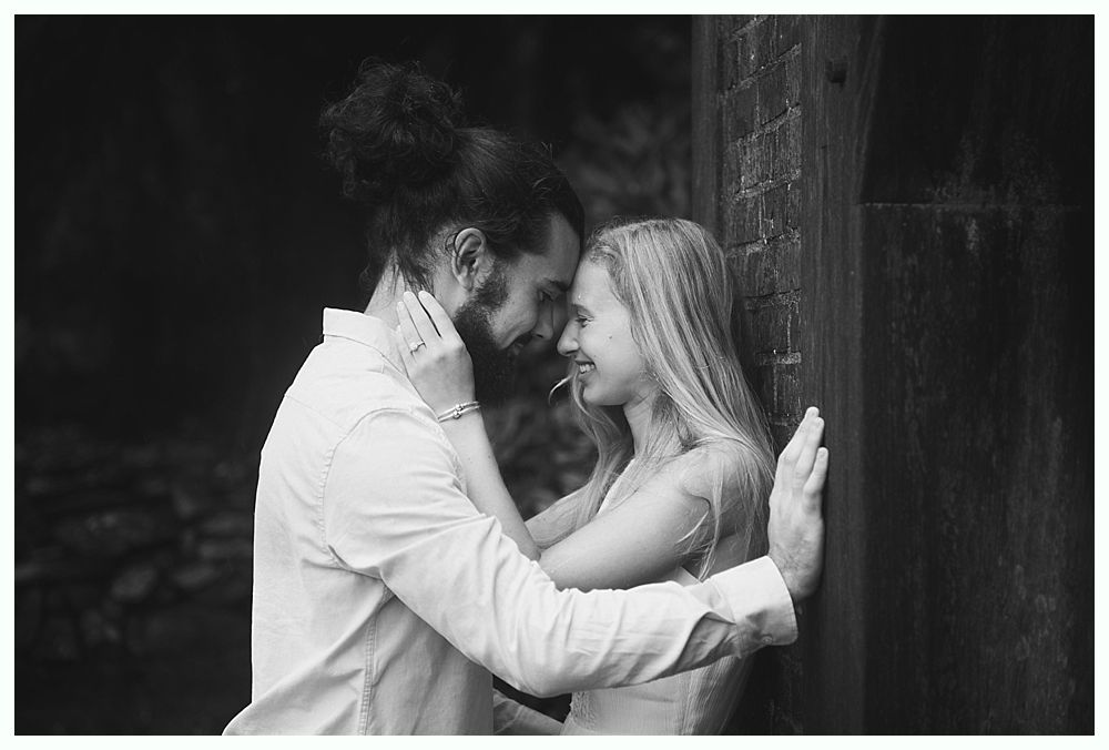 Couple embracing in black and white. Woman smiles, leaning against a wooden wall; man has his face near hers.