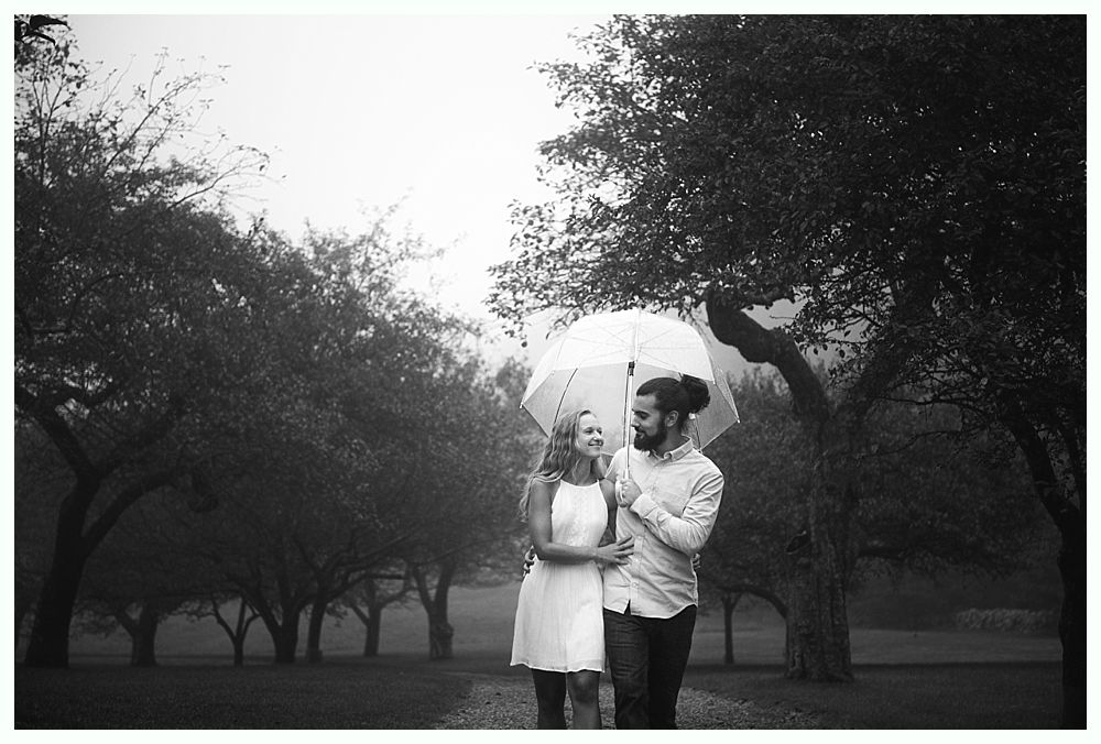 Couple walking under an umbrella in a park, smiling. Trees line the path, overcast sky. Black and white photo.