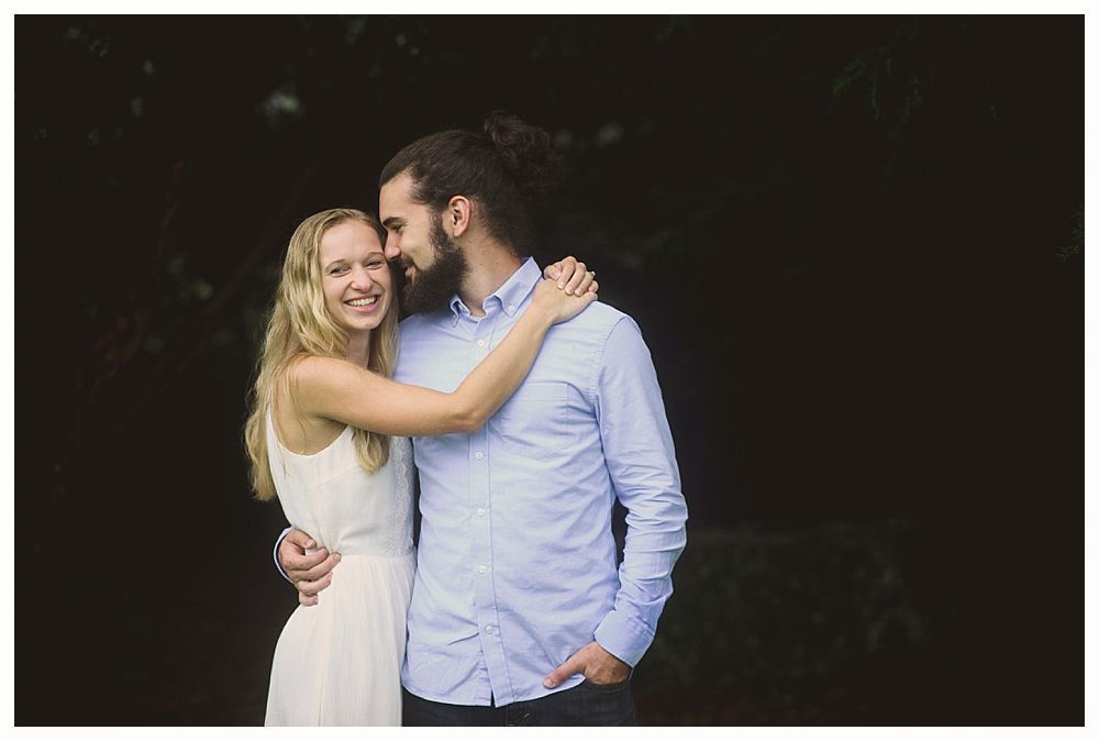 Couple embracing, smiling outdoors; woman in white dress, man in blue shirt, dark background.