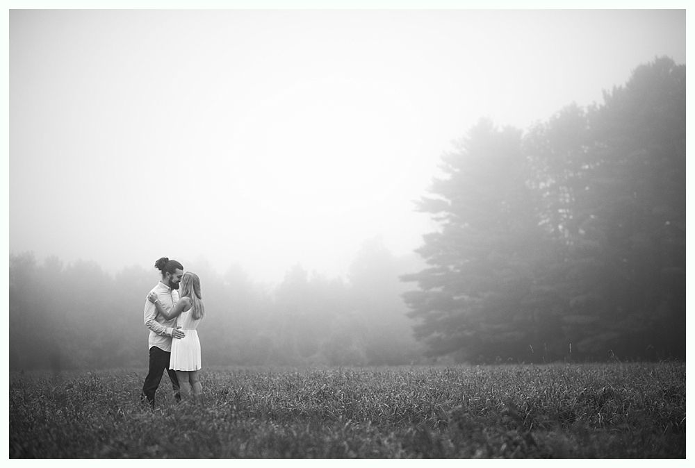 Couple embracing in field, obscured by fog near trees. Black and white photo.