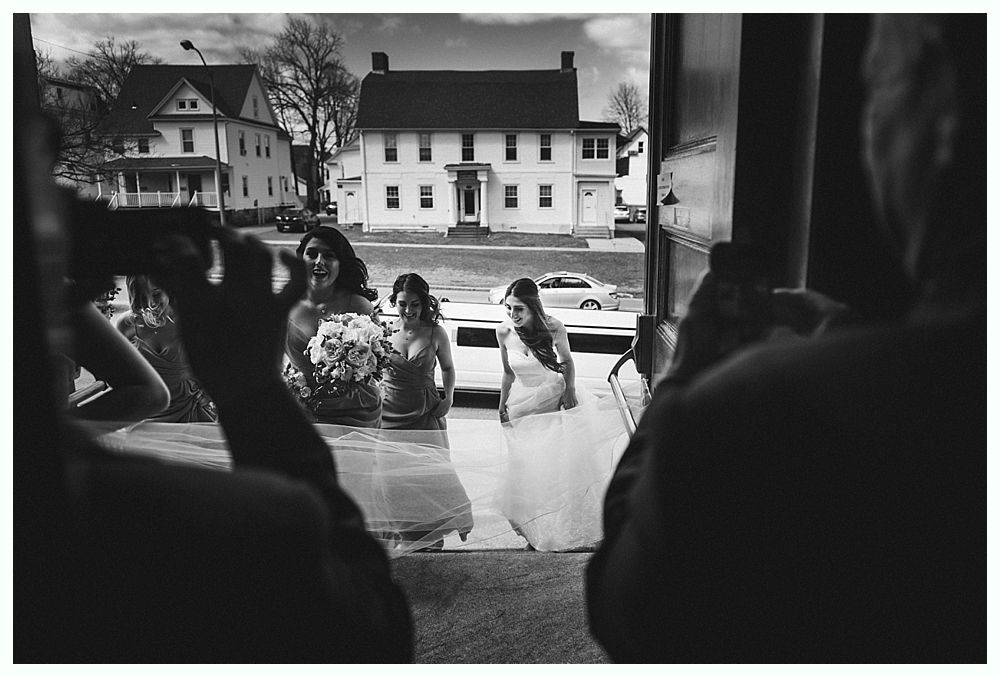 Black and white photo of a bride exiting a building, followed by bridesmaids, with houses and cars in the background.