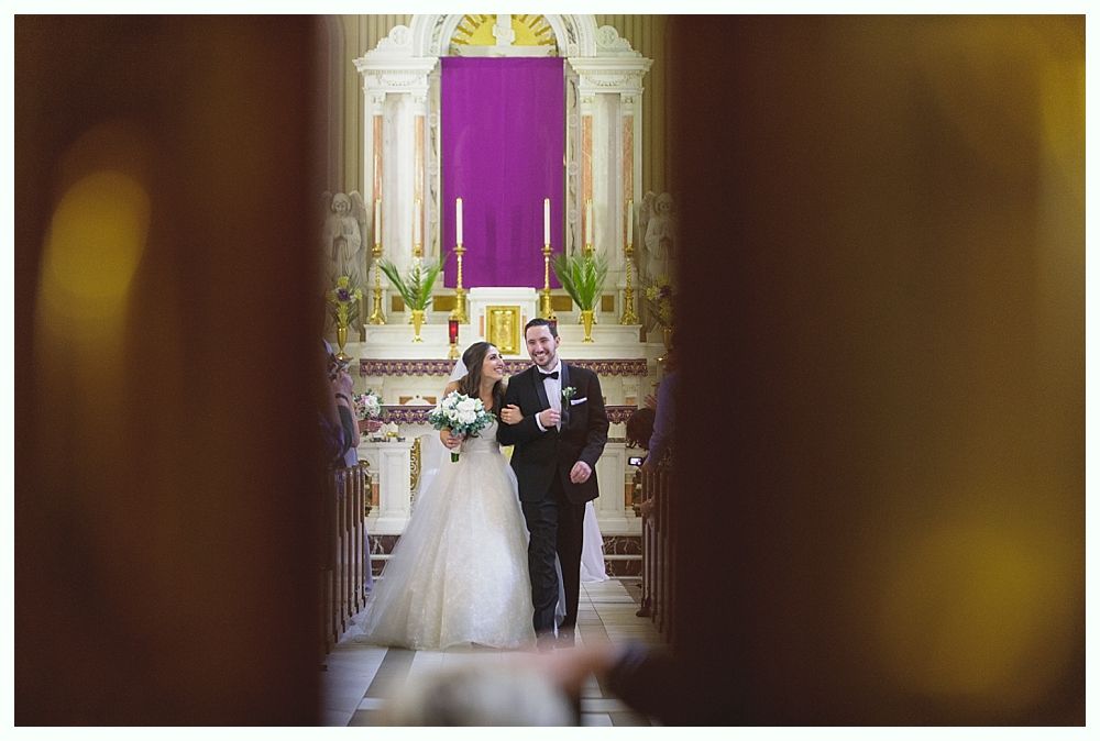Bride and groom walk down aisle in church; bride in white gown, groom in tuxedo.