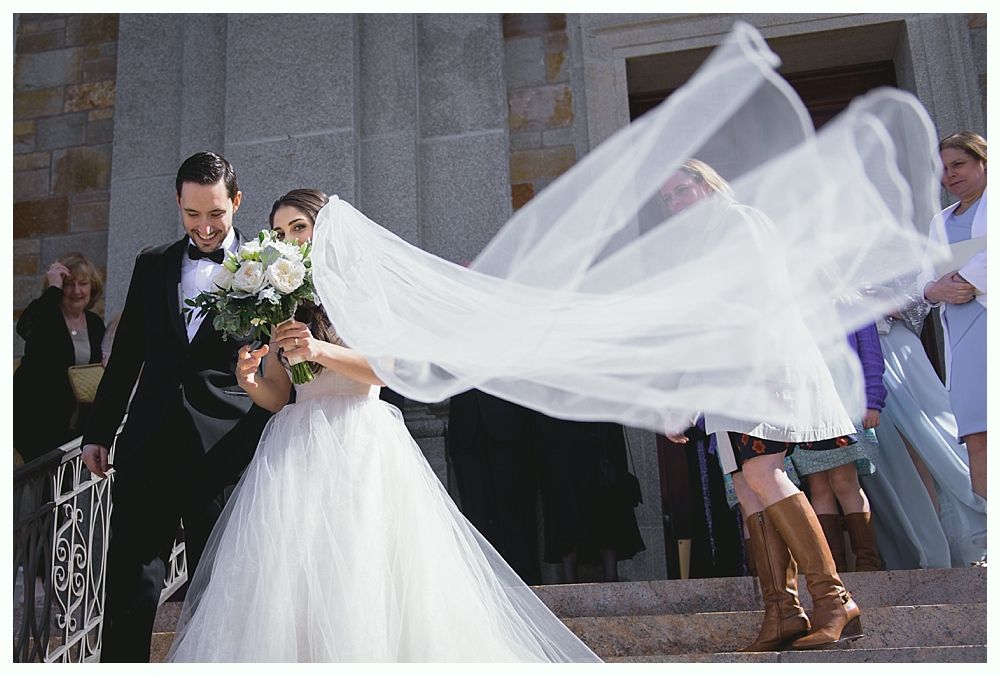 Bride and groom exiting a building, veil blowing in the wind, holding flowers. People in background.
