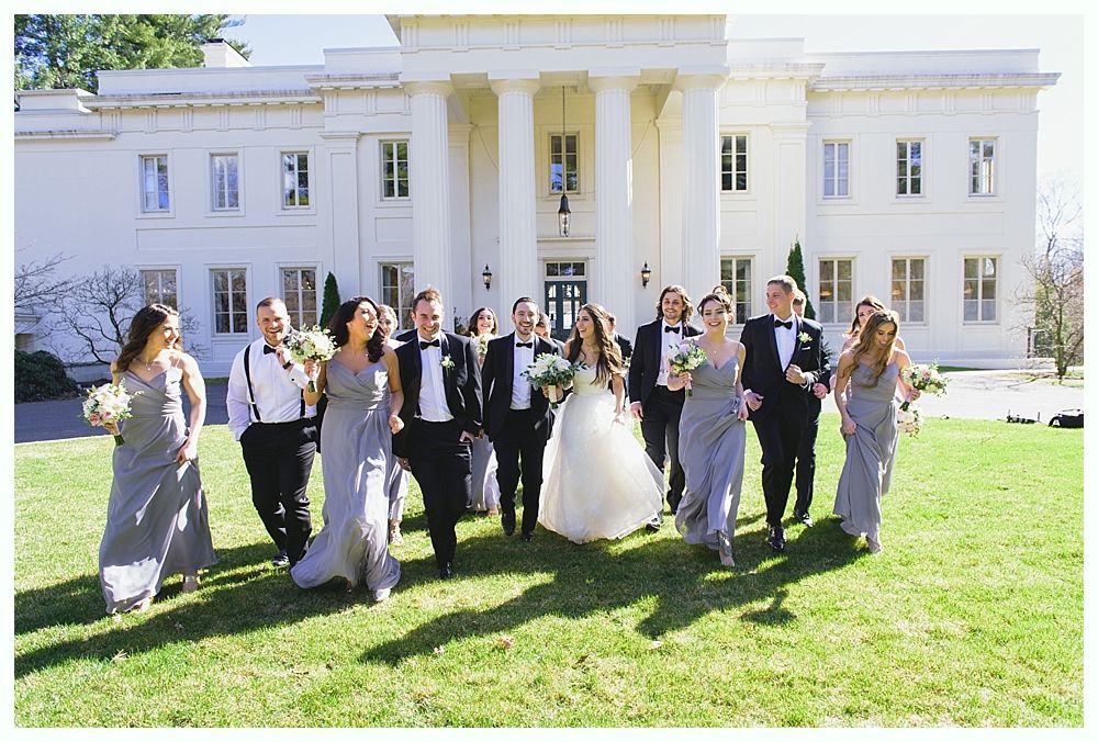 Wedding party on a lawn in front of a white building with columns. Bride and bridesmaids in grey, groomsmen in tuxedos, smiling.