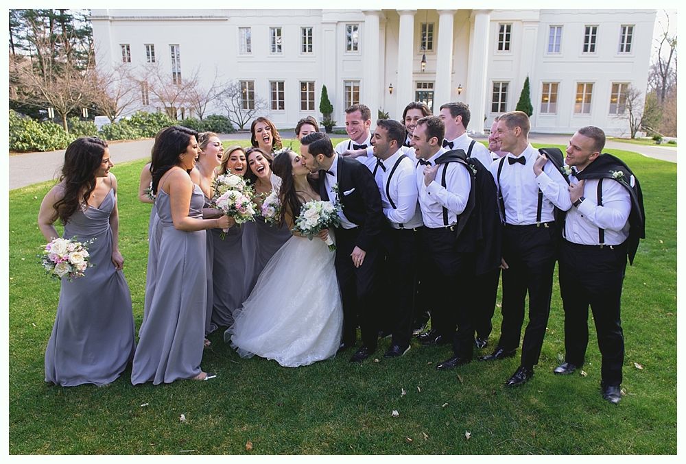 Wedding party outside a white building. Bride and groom surrounded by bridesmaids in gray and groomsmen in black, laughing.