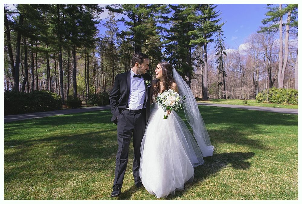 Bride and groom in formal attire, smiling at each other on a sunny grassy lawn with trees in background.