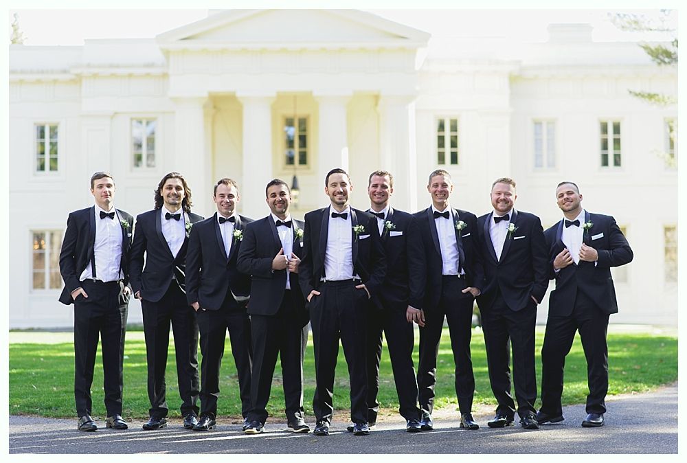 Men in tuxedos pose in front of a white building with columns; all smiling.