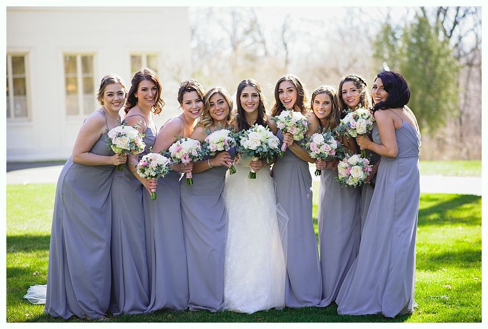 Bride with bridesmaids in blue-gray dresses holding bouquets, standing outdoors on a sunny day.