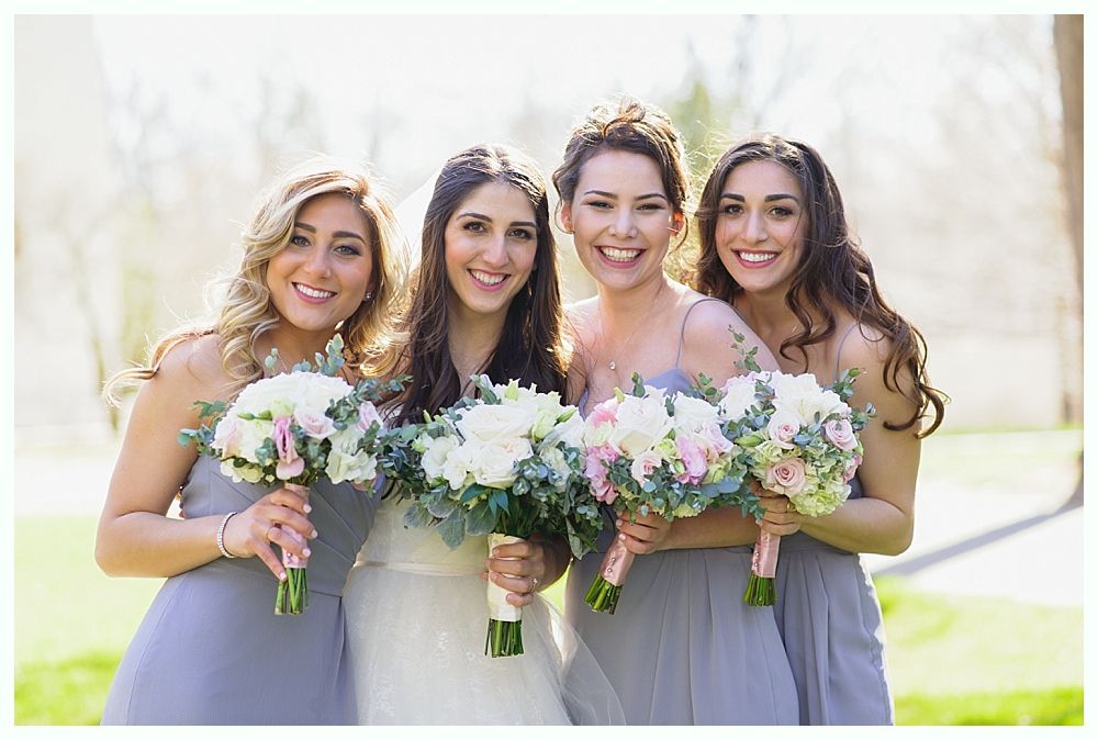 Four women smiling, holding bouquets in a sunny outdoor setting.