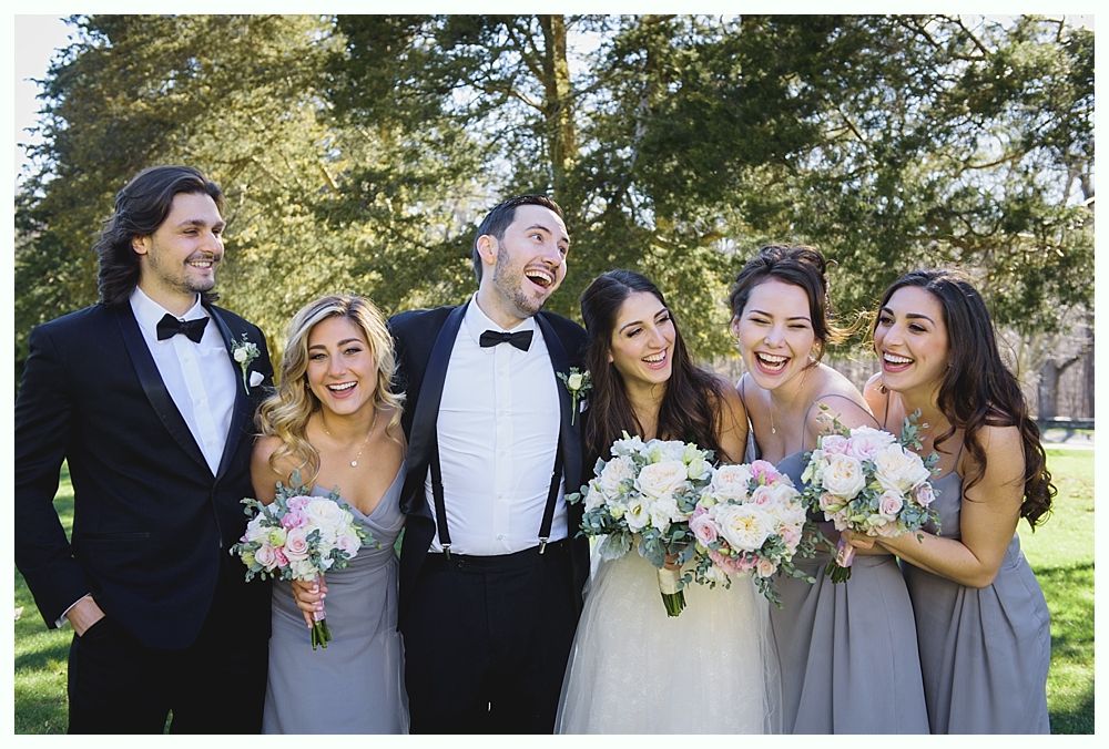 Wedding party laughing outdoors, holding bouquets. The men are in tuxedos, the women in gray dresses.