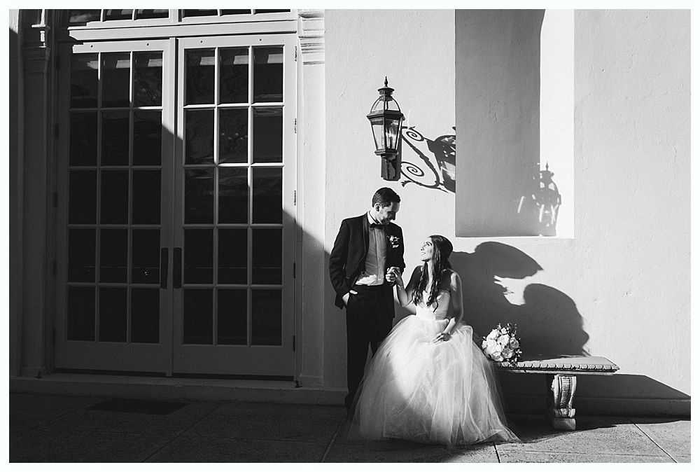 Couple in wedding attire stand near a white building with sunlight casting shadows.