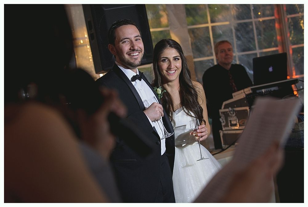 Newlyweds smiling, celebrating at reception. Bride in white dress, groom in tux. DJ in background.