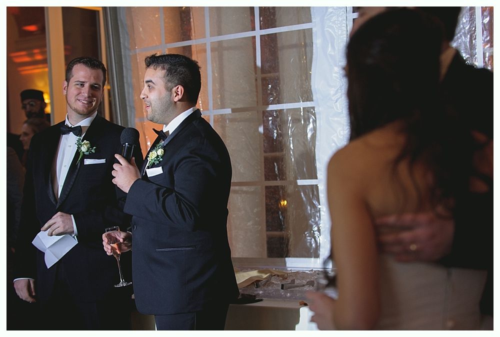 Man in tuxedo giving a speech, other man looks on, couple visible in background.