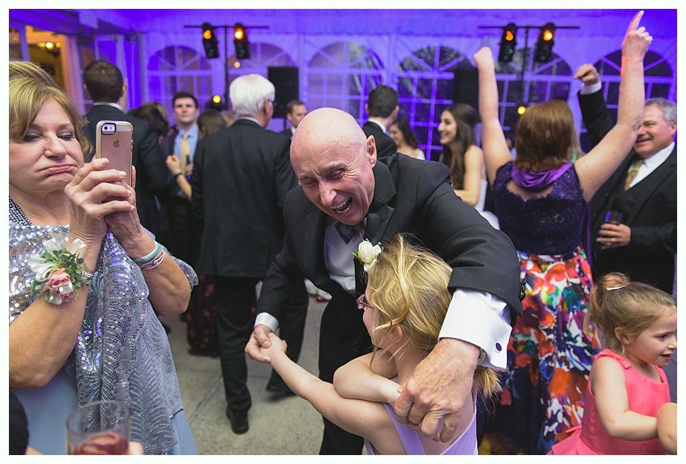 People dancing at an event. A man in a tuxedo dances with a young child; others in formal wear. Purple lighting.