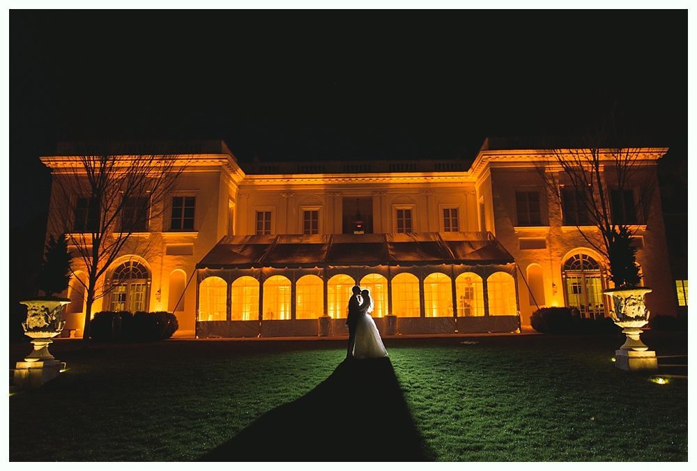 Couple embraces in front of a brightly lit mansion at night. Long shadow in the foreground.