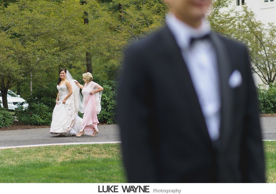 Bride and woman walking toward a man in a tuxedo; trees and a building in the background.