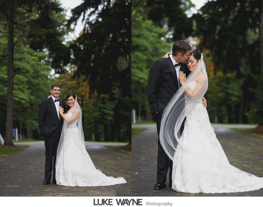 Bride and groom in formal attire, posing outdoors on a tree-lined path. Groom kisses bride's temple.
