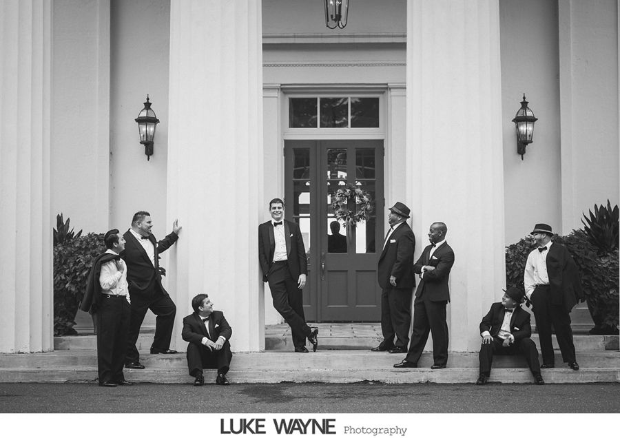 Groomsmen pose outside a building with columns and a red door. Black and white photo. Men wear tuxedos and hats.