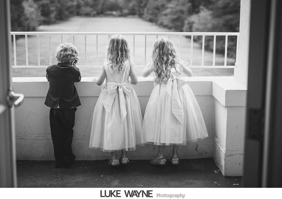 Three young children in formal wear look out over a balcony railing.