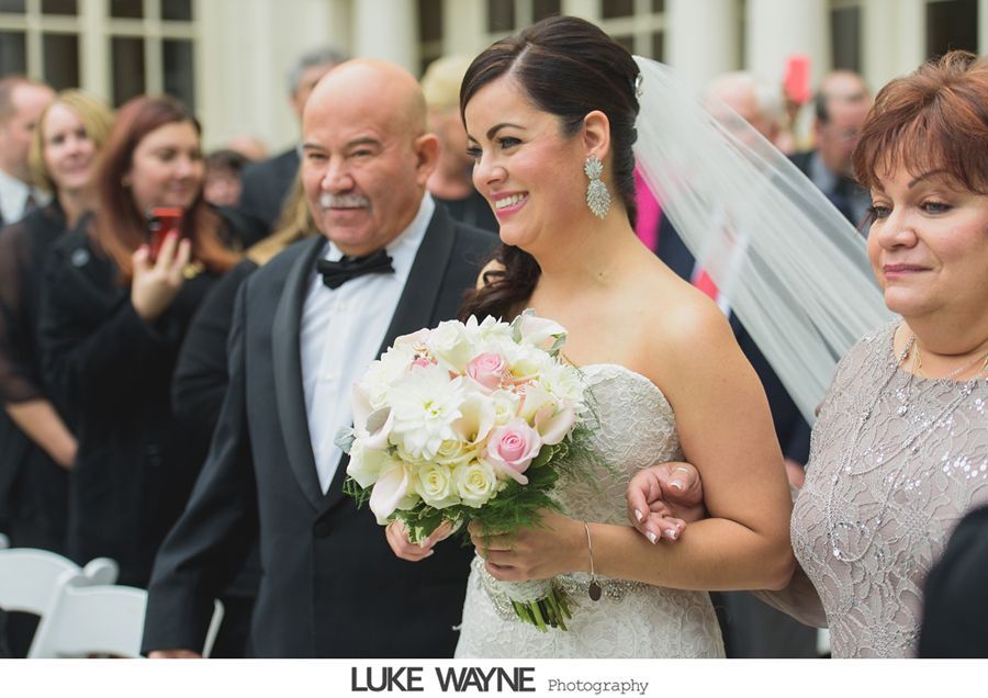 Bride walking with father and mother at a wedding ceremony; smiling, holding flowers, outdoor setting.