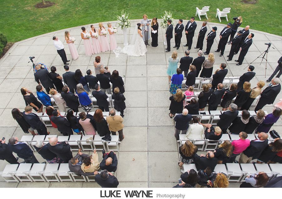 Wedding ceremony, aerial view: Bride, bridesmaids, groom, groomsmen, and seated guests on patio.