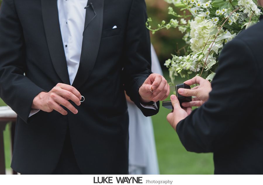 Groom in a tuxedo receiving a ring during an outdoor wedding ceremony.