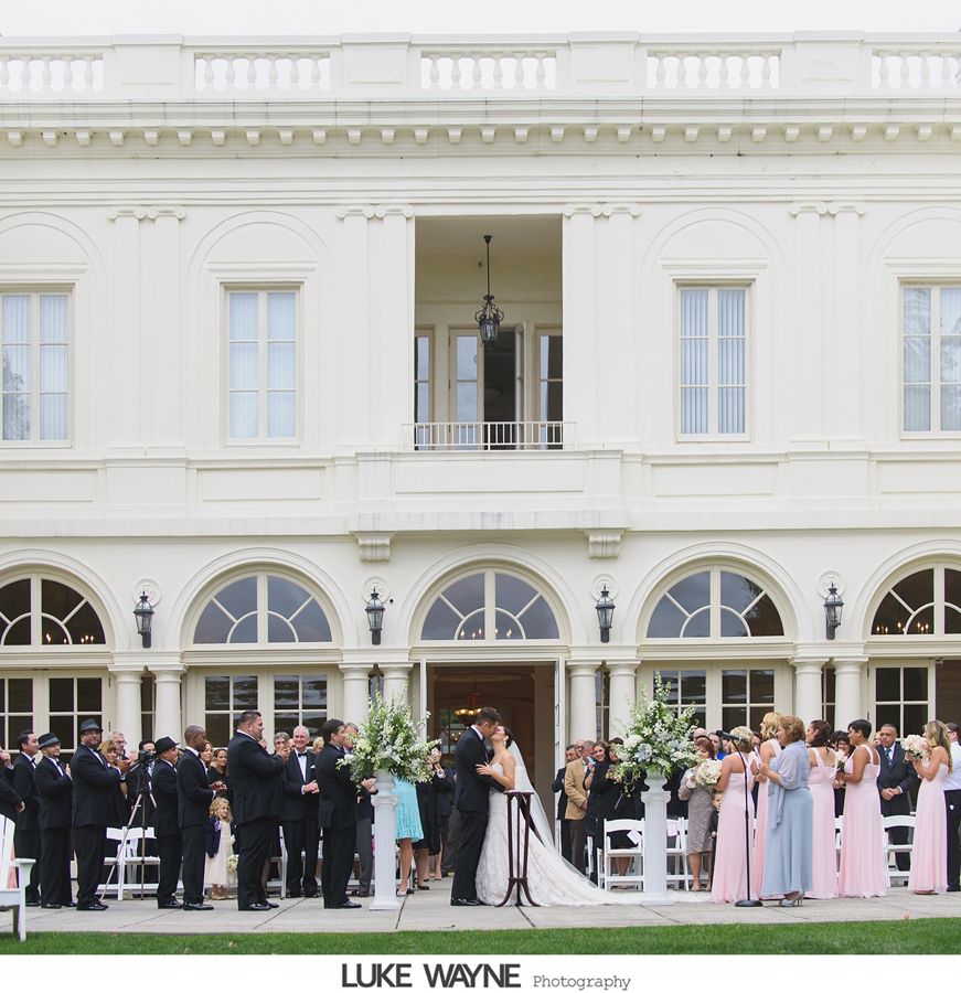 Wedding ceremony outdoors, couple kissing, guests, elegant white building backdrop.