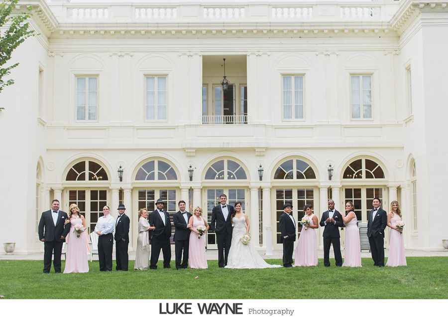 Wedding party stands in front of a white mansion. Bride and groom centered; bridesmaids in pink, groomsmen in black.