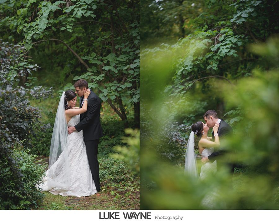 Bride and groom embrace in a lush green forest. The woman wears a white lace gown and veil; the man wears a black suit.