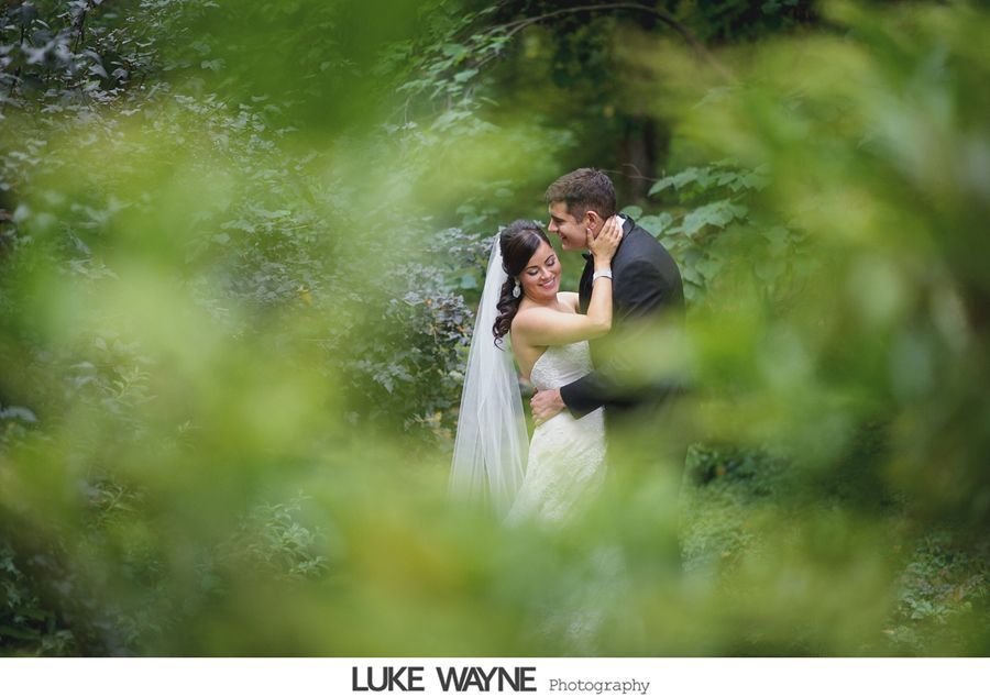 Couple embracing in a forest; the bride wears a white dress and veil; the groom wears a black suit. Green foliage frames them.