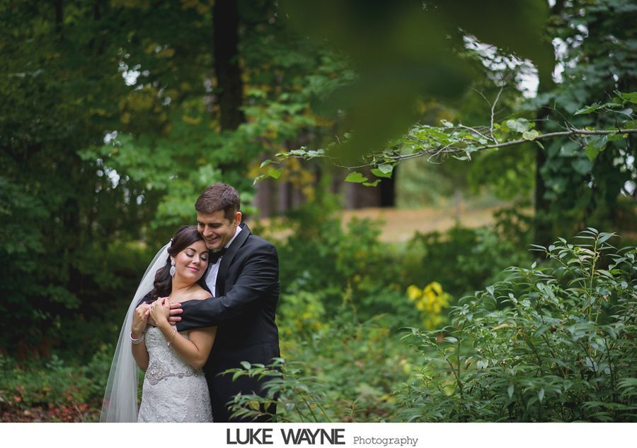 Bride and groom embrace in a forest, the groom's arm around the bride, who smiles. Green foliage surrounds them.