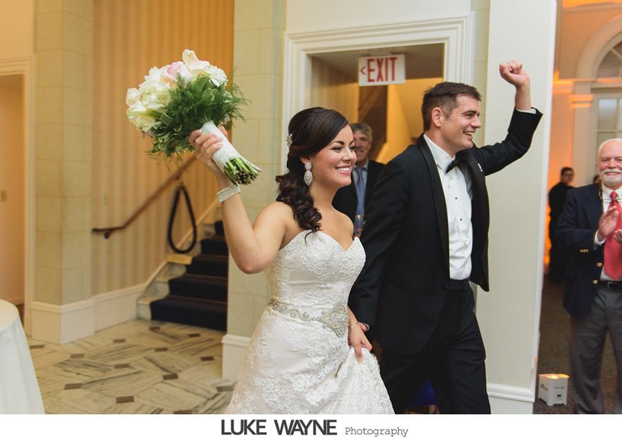 Newly married couple cheering as they enter a reception. Bride holds bouquet, groom raises arm. Formal wear, indoors.