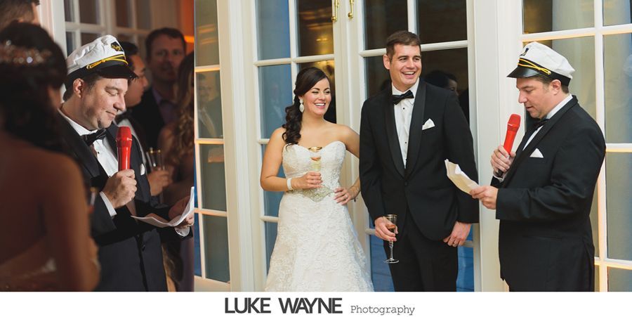 Man in captain's hat speaks at wedding. Bride and groom stand nearby. Window in background.