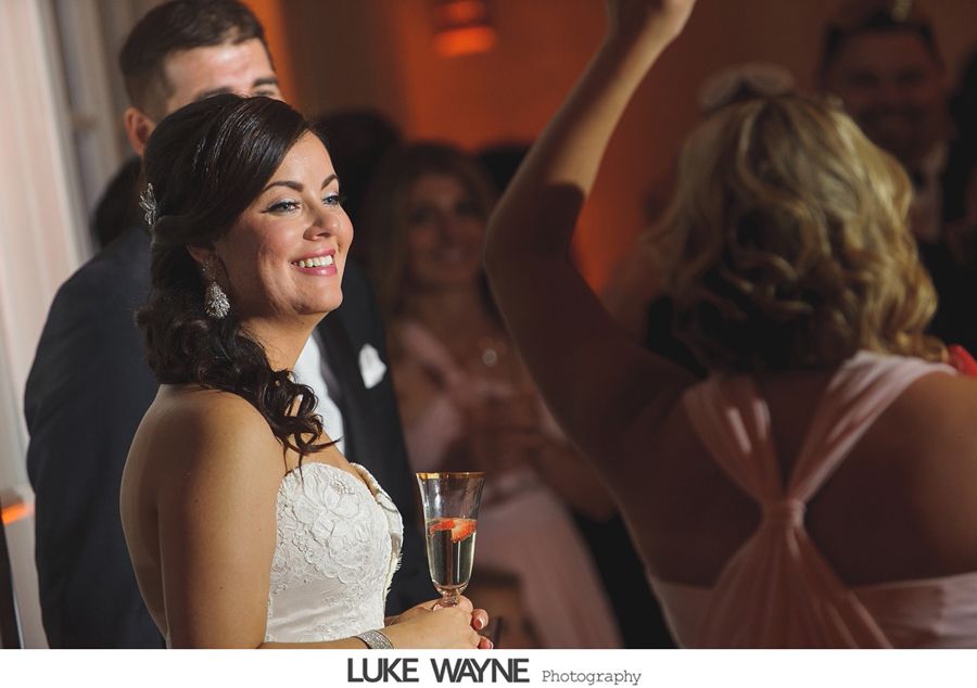 Bride smiling, holding a drink at wedding reception, dancing guests in background. Warm lighting.