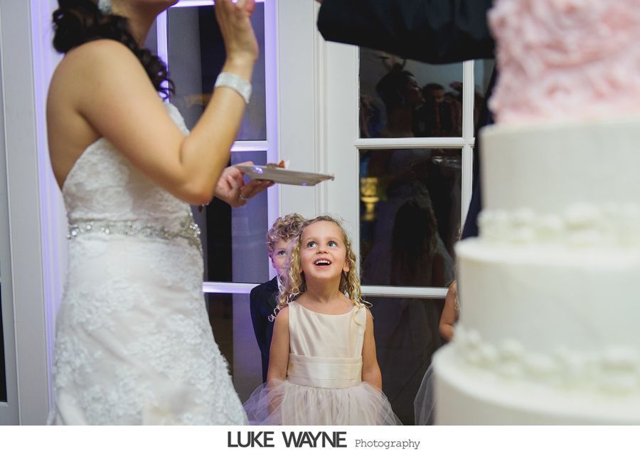 Bride cutting wedding cake, flower girl watches excitedly. Elegant setting with pink and white cake.