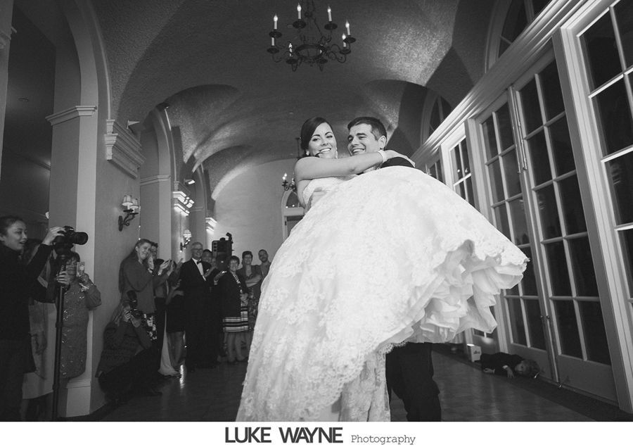 Groom lifts bride in wedding reception. She smiles in a lace gown; guests and a chandelier are visible in the background.
