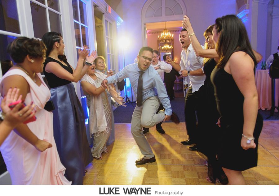 People dancing at a wedding reception on a wooden dance floor. Man in blue shirt kicks his leg up while others cheer.