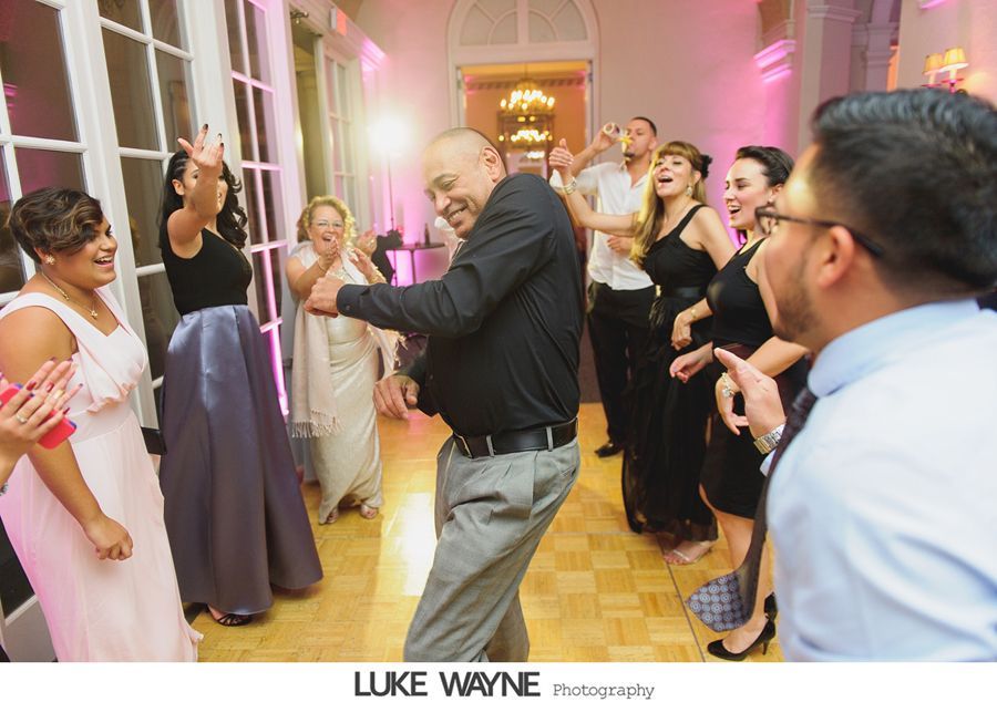 People dancing at a wedding reception. Man in black shirt dances enthusiastically; others clap and smile. Room lit with pink hues.