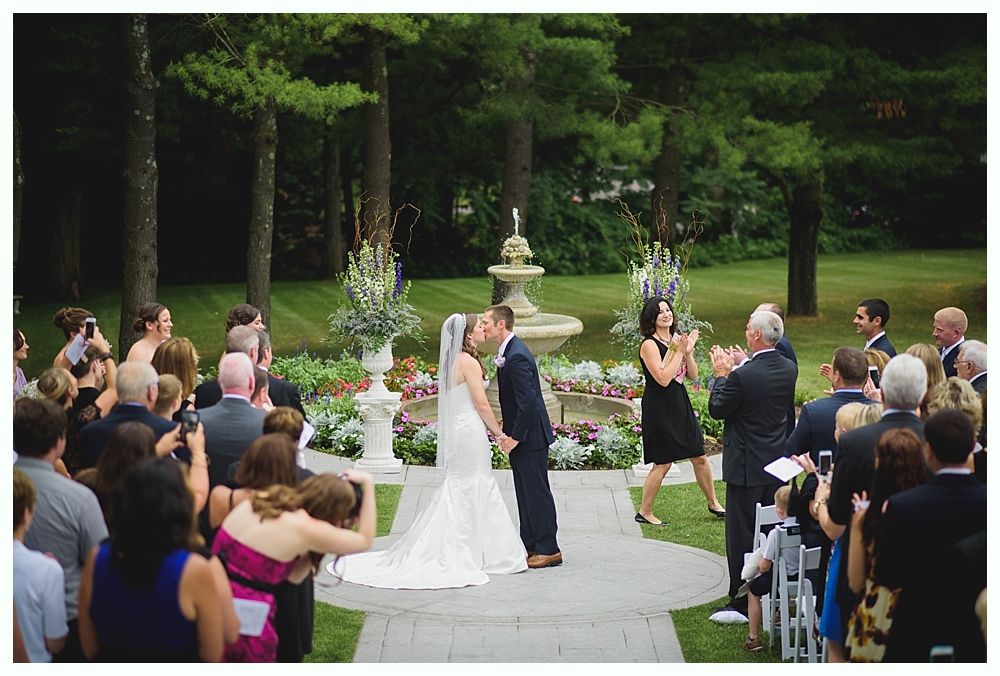 Bride with arms up, making a funny face, groom looking at his hand, cake cutting in background.