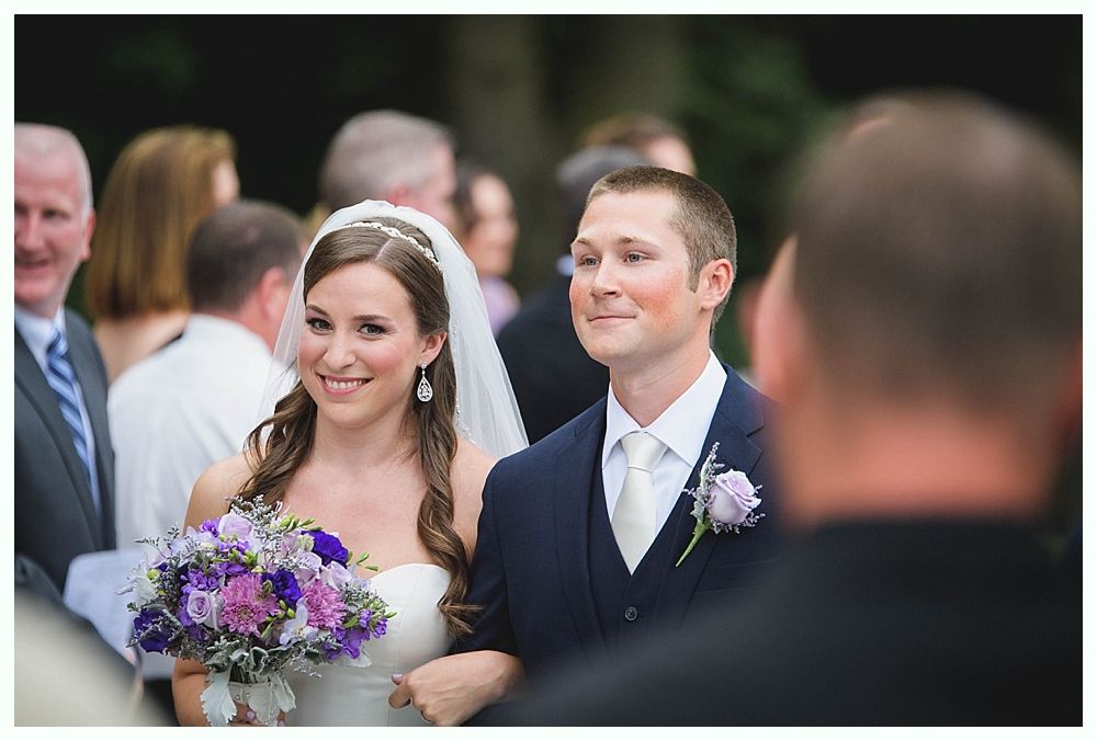 Bride with arms up, making a funny face, groom looking at his hand, cake cutting in background.