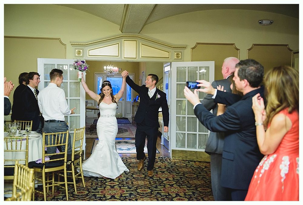 Bride with arms up, making a funny face, groom looking at his hand, cake cutting in background.
