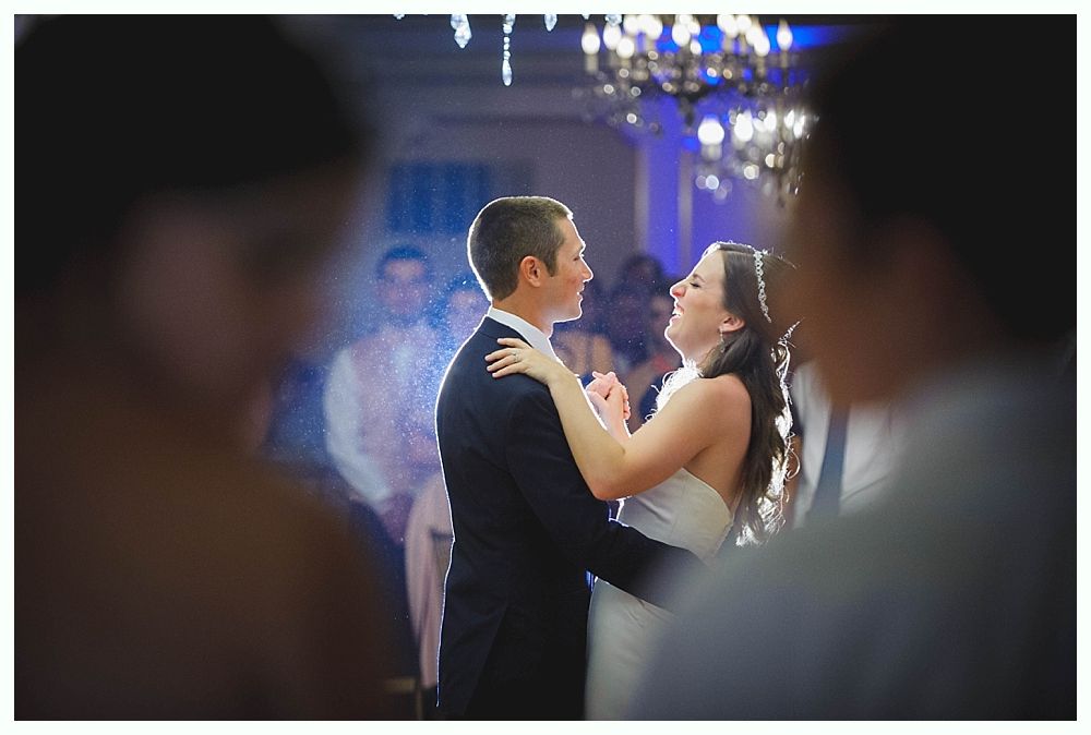 Bride with arms up, making a funny face, groom looking at his hand, cake cutting in background.