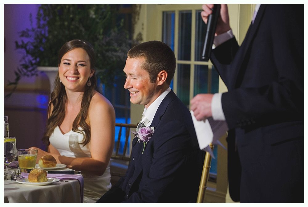 Bride with arms up, making a funny face, groom looking at his hand, cake cutting in background.