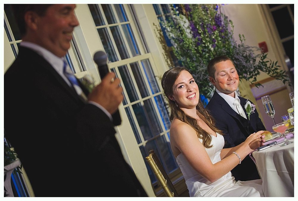 Bride with arms up, making a funny face, groom looking at his hand, cake cutting in background.
